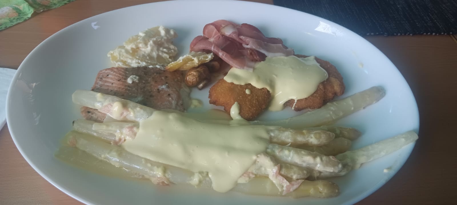 Ein Teller mit Spargel, Schnitzel, Lachs, Schinken und Kartoffeln, bedeckt mit heller Sauce.
